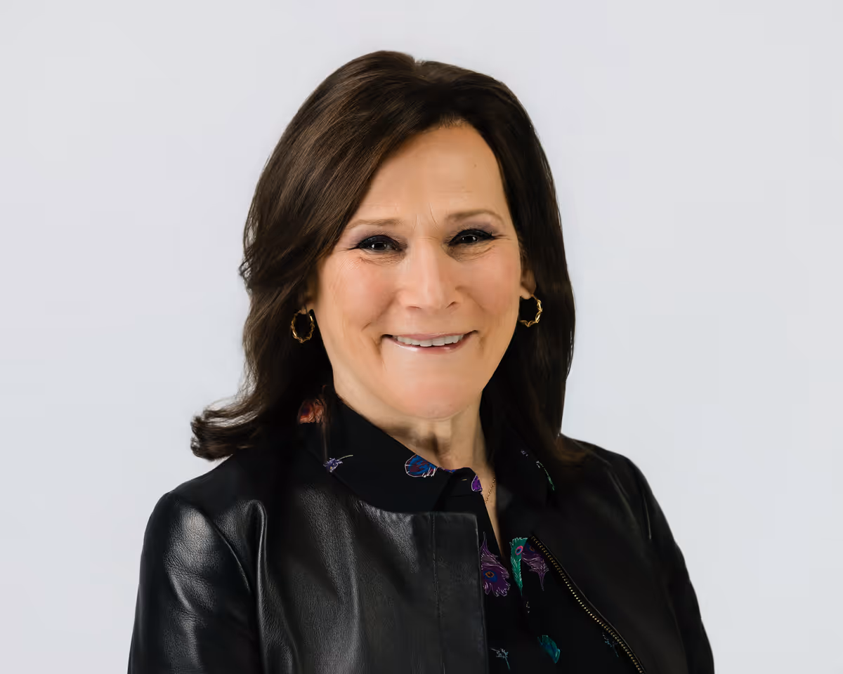 Smiling woman with shoulder-length dark brown hair wearing gold hoop earrings, a black leather jacket, and a patterned blouse against a light gray background.